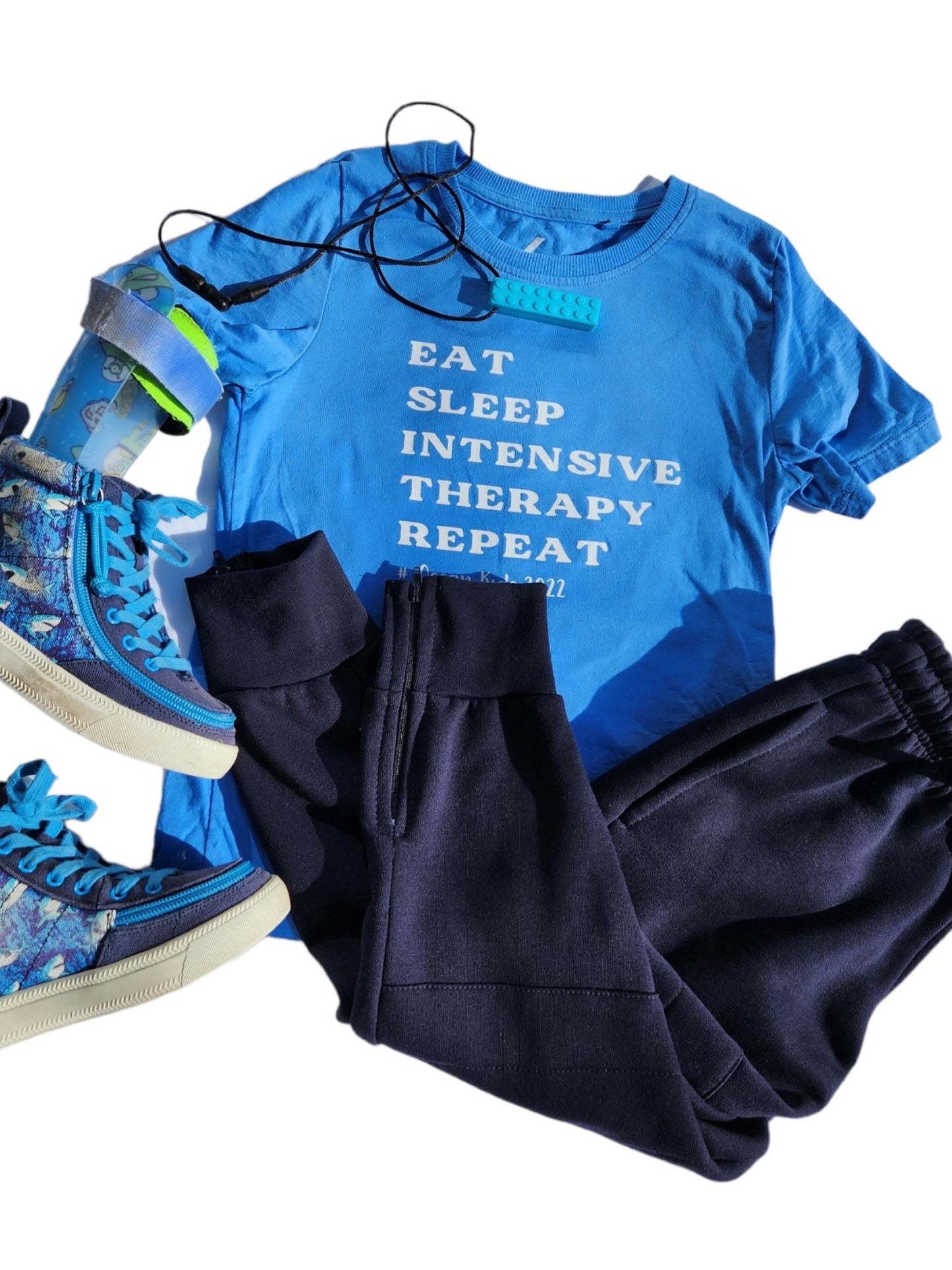 Navy blue cotton fleece track pants with drawstring adjustable waist, knee padding and zips at ankles. Light Blue cotton shirt with writing, blue chewy toy, adaptive shoes and blue ankle foot orthotics (AFO)